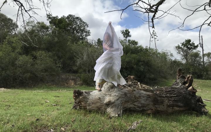 A woman wrapped in white cloth stands on a large piece of deadwood in the middle of a clearing.