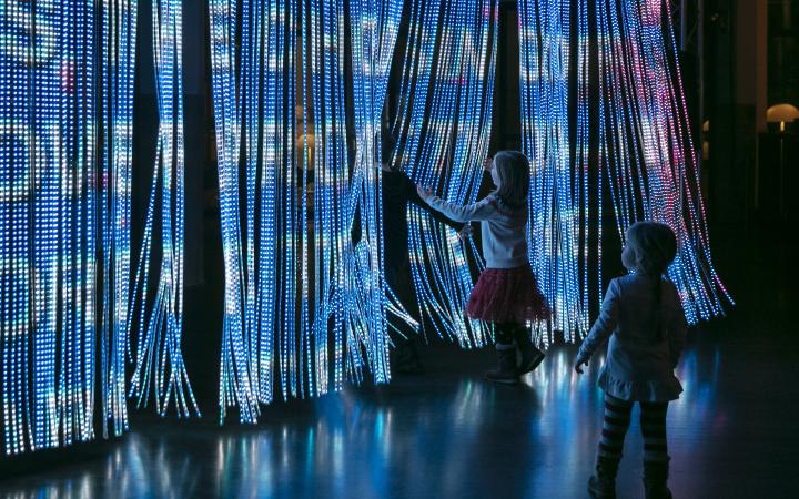Children step through a curtain of light sticks on which many bright colours and lettering are projected.