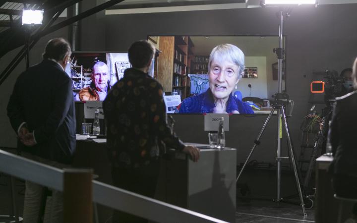 Donna Haraway can be seen on a large screen. Bruno Latour can be seen on another large screen.