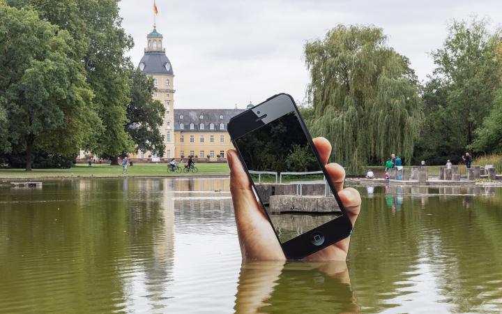 You can see the shore of a lake. A very large hand with a smartphone is sticking out of the lake. The display of the smartphone is a mirror in which the viewers can see themselves on the shore.