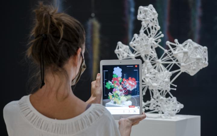 Eine Person steht vor einer weißen 3D-Netzwerk-Skulptur und hält ein Tablet hoch, auf dem mittels Augmented Reality die Skulptur näher untersucht werden kann.