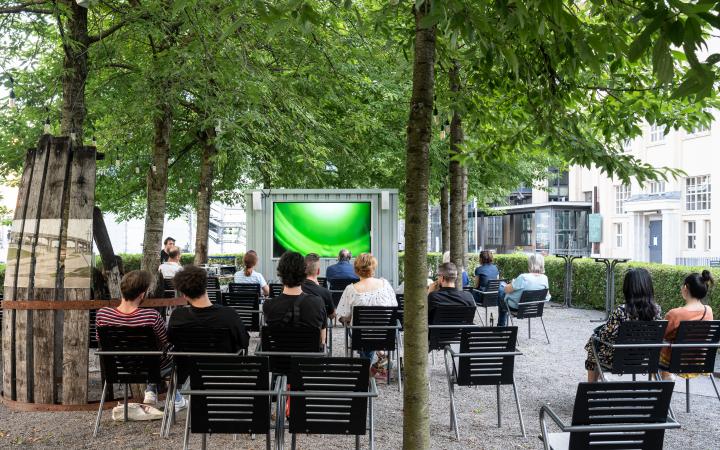 A large screen with people in chairs in front of it outside under green trees.
