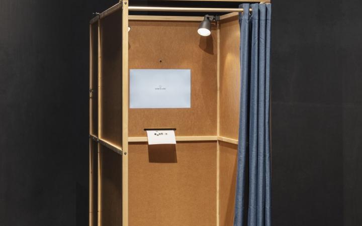 Das Bild zeigt eine Wahlkabine aus Holz mit einem blauem Vorhang. Sie ist Teil des Gesamtkunstwerks »Smile to Vote – Political Physiognomy Analytics«, ein künstlerisch-wissenschaftliches Forschungsprojekt, das die Auswirkungen von KI-basierten biometrisch