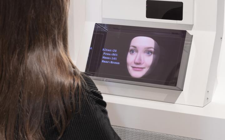 A visitor can be seen. She is seated with her back turned and is interacting with the media installation. She is talking to a female artificial intelligence that is facing her.