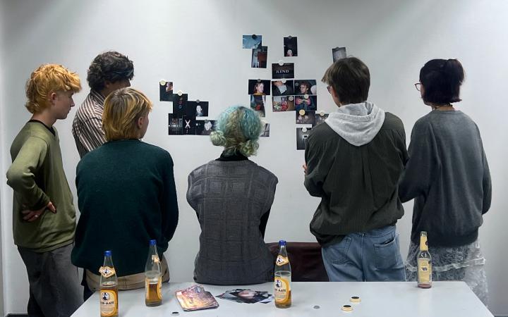 Fünf junge Kunststipendiat:innen sind von hinten zu sehen, sie stehen vor einer weißen Wand an der Fotos hängen