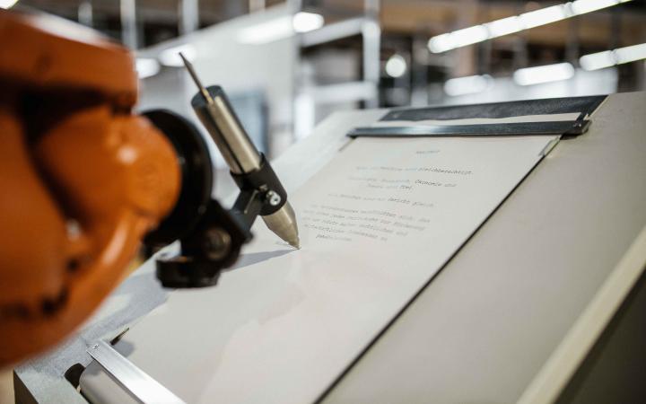 An orange robot arm writes with a silver pen on a sheet of paper