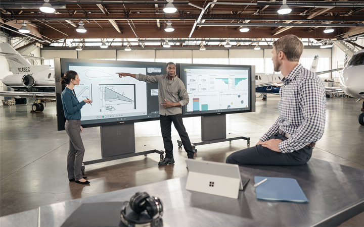Drei Menschen arbeiten an digitalen Bildschirmen in einer Halle mit Flugzeugen
