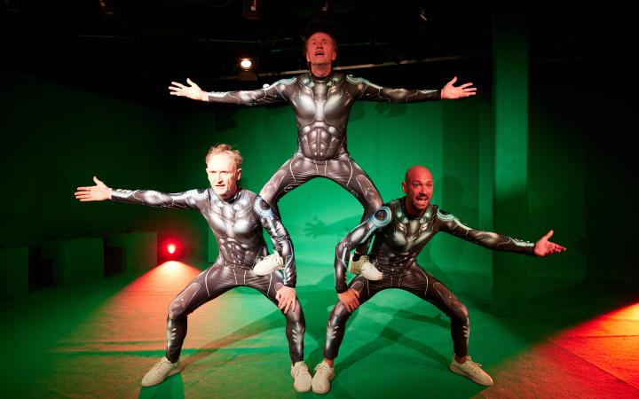 Drei Männer in futuristischen Rüstungen machen eine menschliche Pyramide in einem Green-Screen-Studio