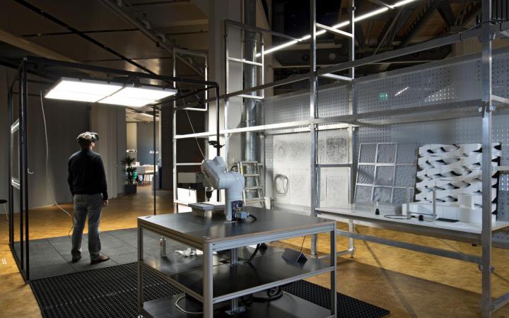 Installation mit einem Roboter und gefrästen Elementen in grauen Regalen, neben welchen ein Mann mit einer VR-Brille steht