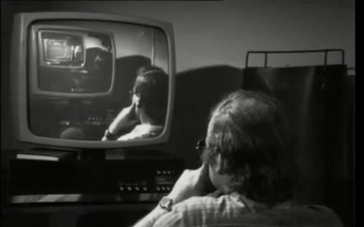 You can see a black and white picture on which an elderly man sits in front of a tube screen on whose screen you can see a young man standing in front of an old television.