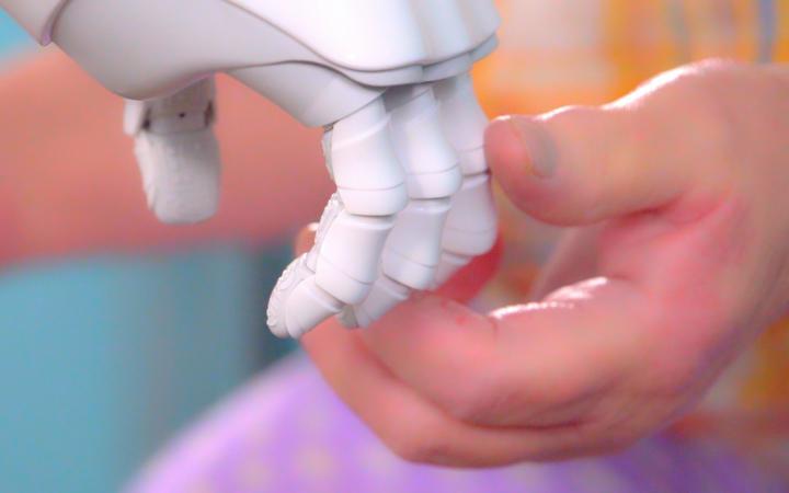 Es ist ein animiertes Bild welches aussieht wie der Screenshot eines Videos. Auf dem Bild erkennt man eine virtuelle Roboterhand die eine virtuelle menschliche Hand and den Fingerspitzen hält.