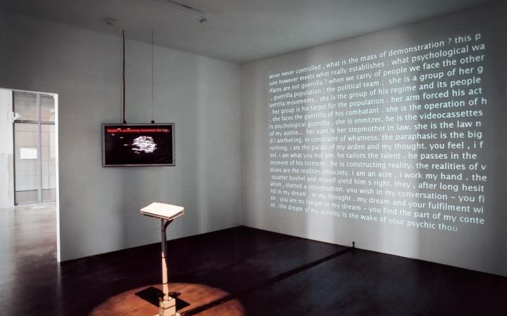 In einem Raum steht eine Tastatur. An einer Wand hängt ein Bildschirm, auf eine andere Wand wird Text projiziert. 