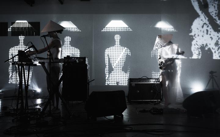 The Duo group A is performing live on stage, in the background there are visuals with abstract figures wearing the traditional asian cone-hat