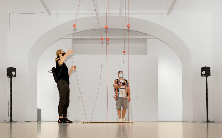 A man and a woman stand in front of a work of art consisting of ropes stretched between the ceiling and the floor. The visitors pull the ropes apart.
