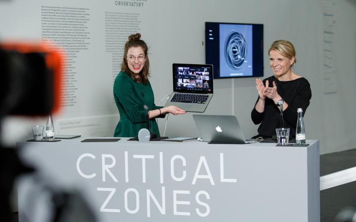 Two women stand behind a desk. One woman is holding an open laptop, beaming with joy.