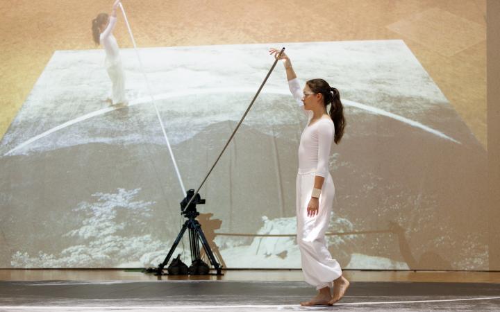 Performance von Ulrike Rosenbachs »Die einsame Spaziergängerin«, dir Künstlerin, ganz in weiß gekleidet hält eine Stange fest und läuft im Halbkreis einen Weg ab, während sie gefilmt wird.