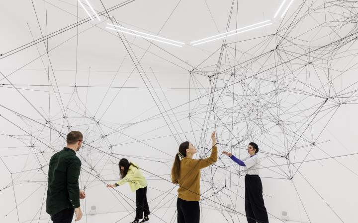You can see a large net of threads in a white room. Several people are standing in this space and touching these threads.