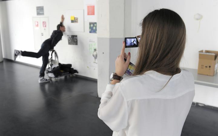 Eine junge Frau fotografiert einen für das Bild posenden Mann, der so tut, als ob er mit einem Rollwagen gegen die Wand fährt.