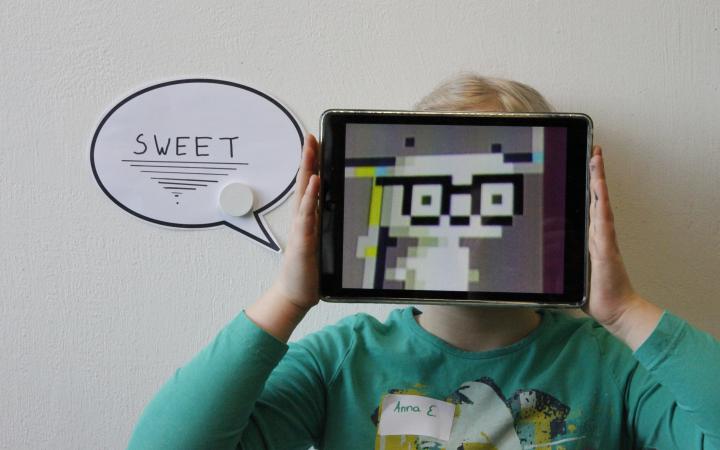 Performance of a girl, that is holding an iPad in front of her face. The display shows a creature buid of pixels. Next to her, the word "sweet" is written in a speech bubble.