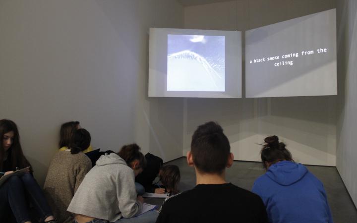 Jugendliche sitzen vor zwei großen Projektionsflächen, auf denen ein Bild und die Zeile »a black smoke coming from the ceiling« zu sehen sind.