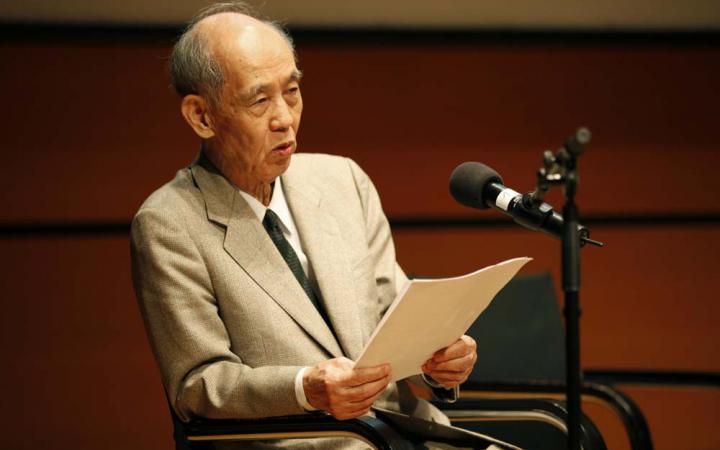 Hiroshi Kawano bei seinem Vortrag. Er trägt einen hellen Anzug, in den Händen hält er sein Manuskript.