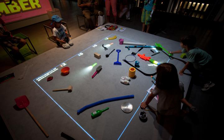 Kinder sitzen vor einer Spielfläche auf der Gegenstände liegen und virtuelle Autos fahren