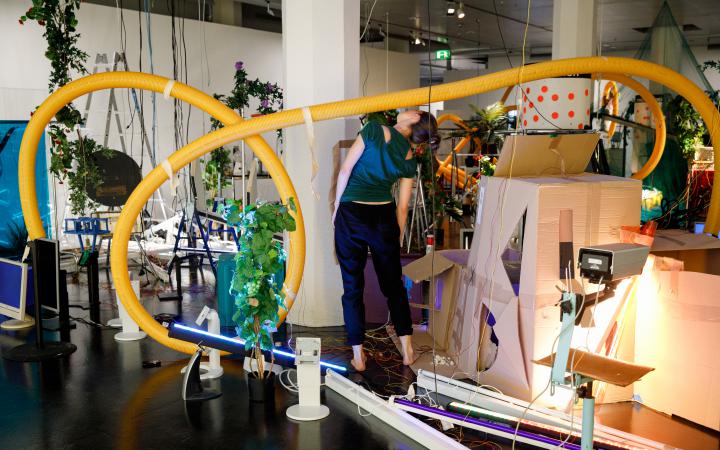 The photo shows an insight into the performance of the opening of »Edge of Now« in the midst of an artinalllation of plants, cardboard and plastic pipes.