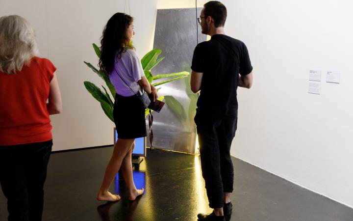 Das Foto zeigt eine barfüßige Besucherin und einen schwarz gekleideten Besucher vor einer an der Wand angelehnten Installation neben der eine Pflanze steht