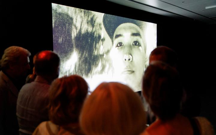 Das Bild zeigt eine Menschenmenge vor einem riesigen Bildschirm auf dem gerade ein asiatischer junger Mann mit Cappy zu sehen ist.