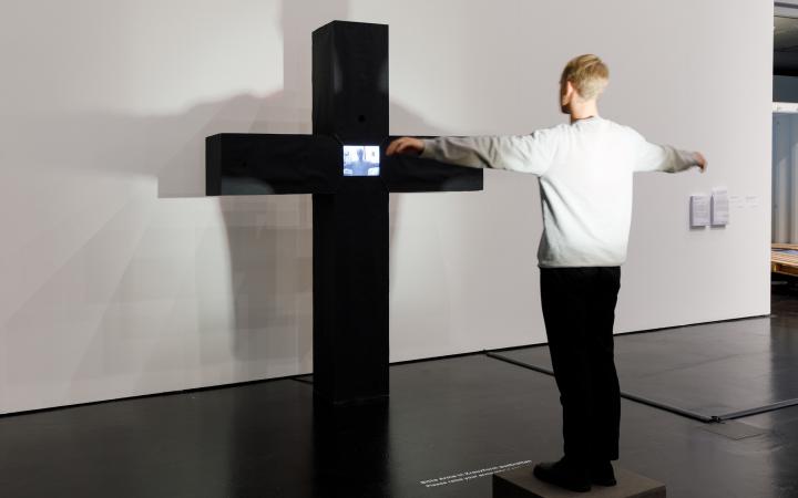 Peter Weibel's interactive video culture: A massive black cross. Where the struts of the cross meet is a monitor. On the monitor you can see the person standing in front of the cross, stretching out his arms as if he himself was hanging there.