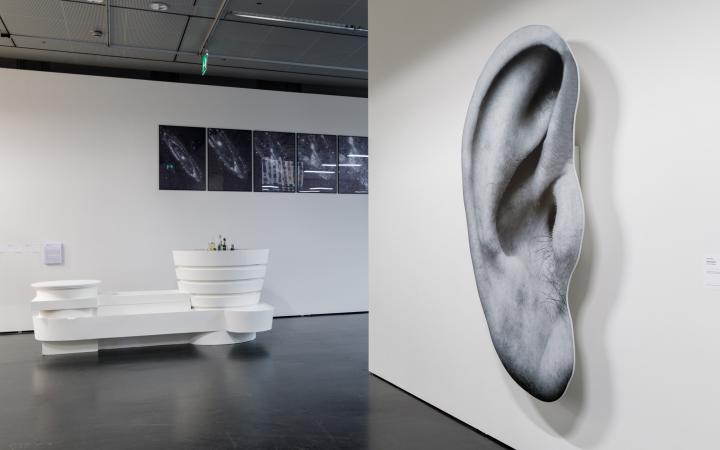 Links eine weiße Installation, die aussieht wie eine futuristische Bar. Im Hintergrund schwarz-weiß Bilder des Universums. Auf der rchten Bildhälfte eine Wand mit einem übergroßen Druck eines Ohres.