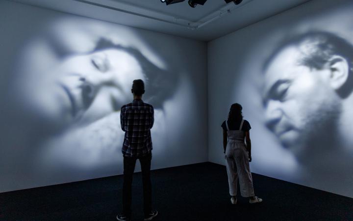 In a dark room, videos of two sleeping people are projected onto the wall. In front of them are two viewers.