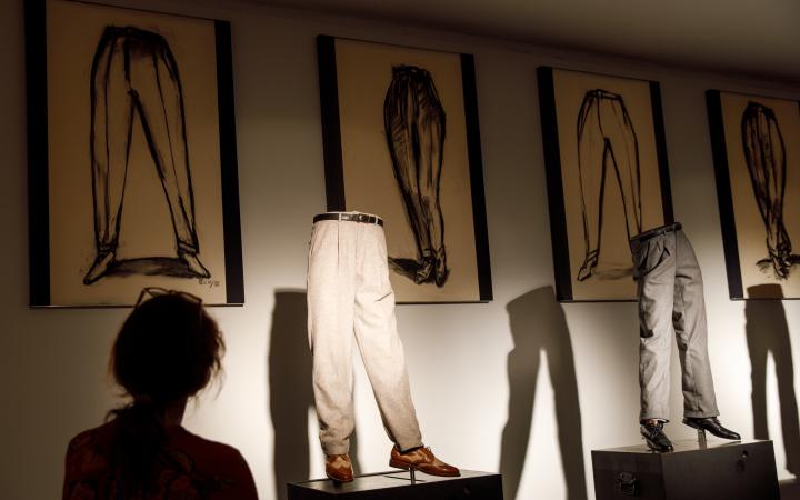 Photo of an art installation: trousers with shoes on a kind of catwalk, framed drawings in the background.