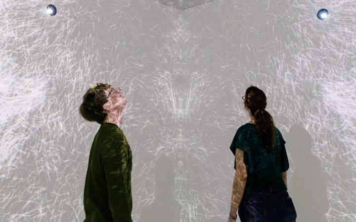 Visitors in a room with video projections on walls and ceiling and a mirrored floor.