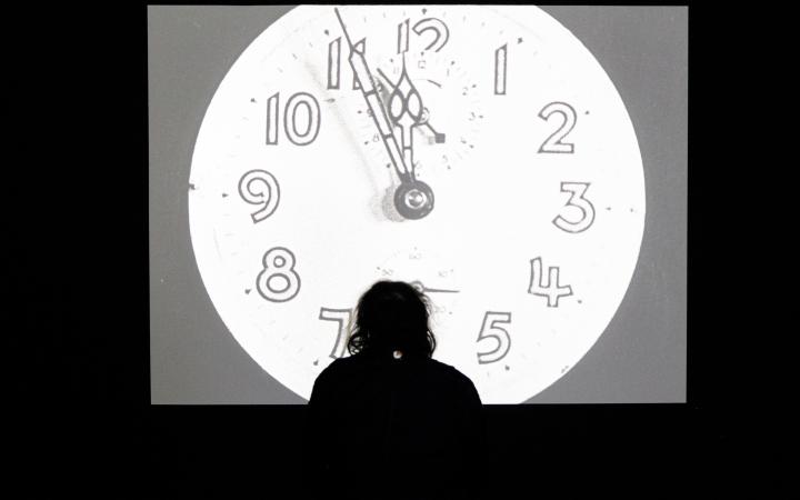 The photo shows the silhouette of a person watching a black and white film on a large screen. On the screen, an analogue clock strikes twelve. The light of the screen falls a round blurred light on the floor.