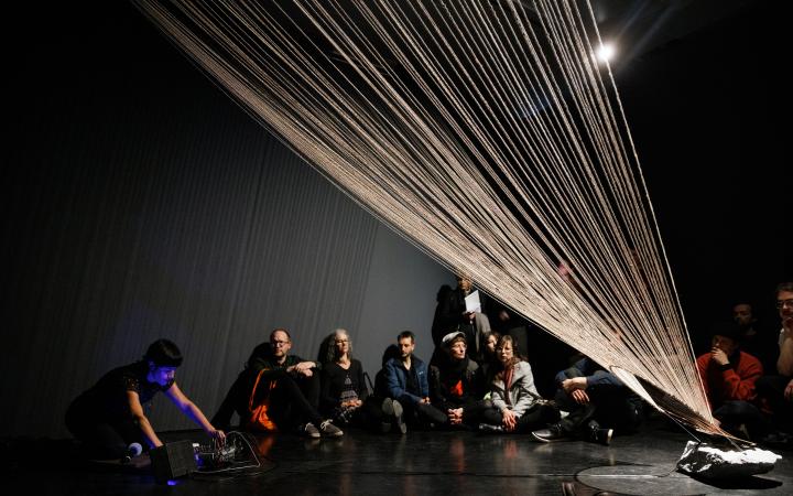 You can see a large, fanned net of threads that reaches up to the ceiling. In the background, several people are sitting against a wall.