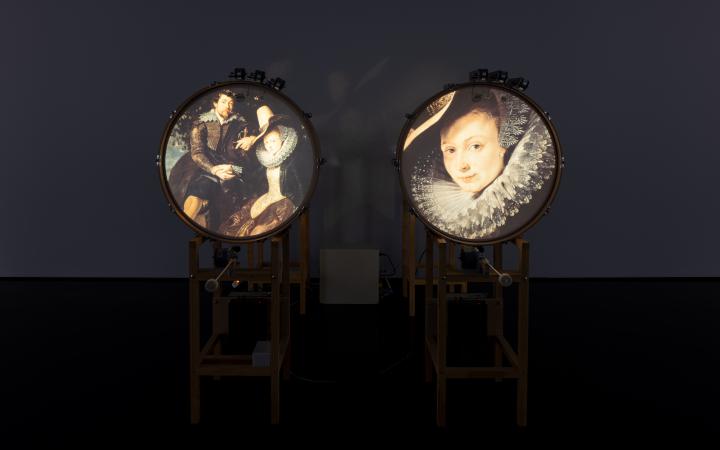 Zu sehen sind zwei stehende Trommeln, welche jeweils ein Bild auf der runden Fläche zeigen. Links ein altertümliches Ehepaar und rechts das Porträt einer Frau. 