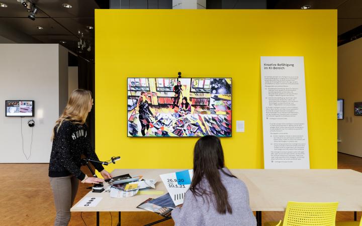 Zu sehen sind zwei Personen, eine stehend, eine sitzend, an einem Tisch. An der Wand vor ihnen hängt ein Bildschirm mit einer Kamera. Auf dem Bildschirm ist in abstrakter und künstlerischer abgebildet, was die Kamera aufzeichnet.