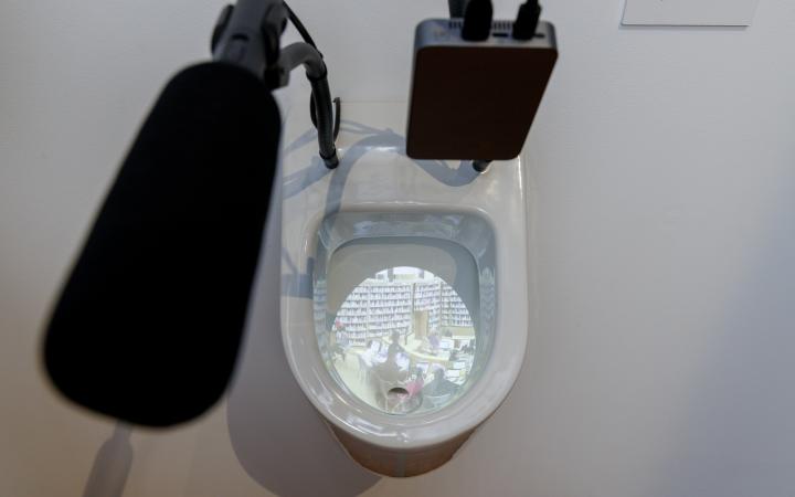 You can see a toilet bowl photographed from above. In it there is a projection. A microphone is fixed on the toilet.
