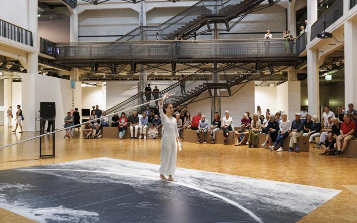Performance von Ulrike Rosenbachs »Die einsame Spaziergängerin«, dir Künstlerin, ganz in weiß gekleidet hält eine Stange fest und läuft im Halbkreis einen Weg ab, während sie gefilmt wird. Im Hintergrund sieht man ein großes Publikum.