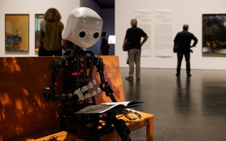Zu sehen ist ein Roboter, dessen Aufbau dem Korpus des Menschen gleicht. Er sitzt auf einer Bank.