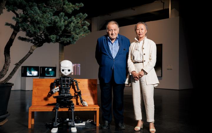 Zu sehen ist ein Roboter, dessen Aufbau dem Korpus des Menschen gleicht. Er sitzt auf einer Bank. Recht daneben steht Peter Weibel zusammen mit Soun-Gui Kim.