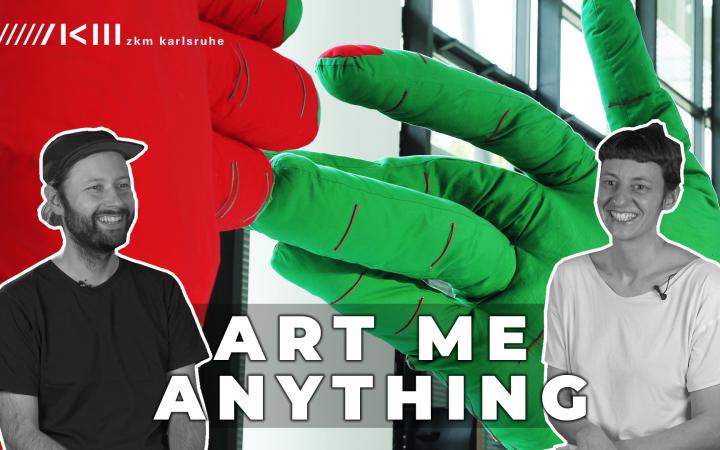 Photo of a huge green and a red fabric hand that touch each other. In front of it the writing "Art me anything" and two people.
