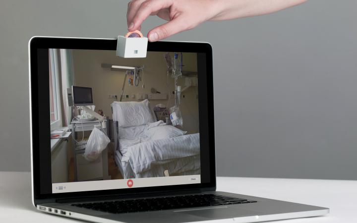 Photo of »Object A – Un-hacking Webcams« of the work »Accessories for the Paranoid«, laptop with a small device with a dial in front of the webcam, on which a hand turns. On the screen you can see a hospital bed room.