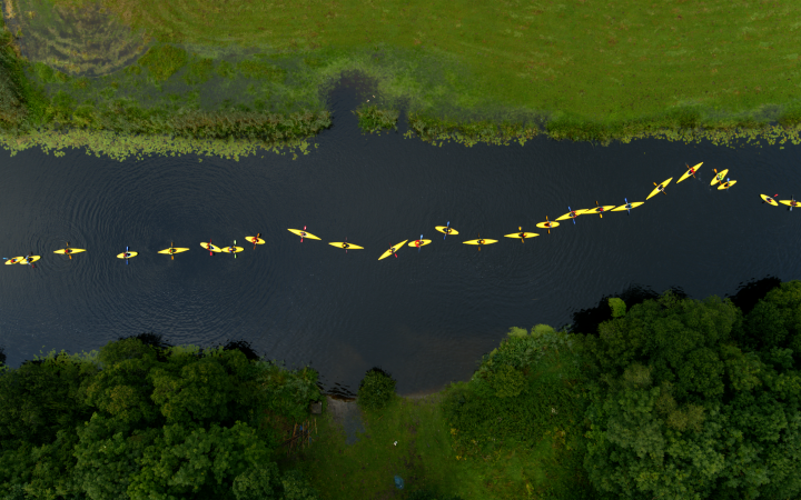 You can see a green landscape with a river meandering through it. The picture shows a bird's eye view of nine people in yellow rain jackets and yellow canoes paddling along the river.