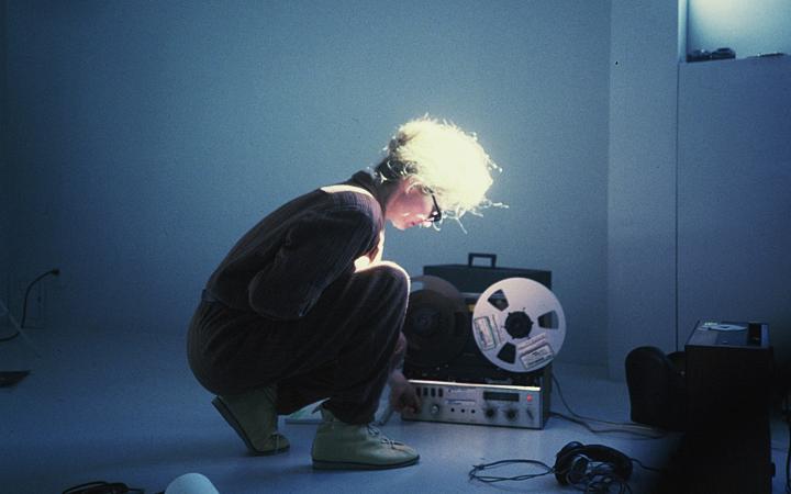 In the picture, a woman is sitting in a crouching position and adjusting something on a receiver that is standing diagonally next to her. Around her are some technical objects on the floor and she herself has sunglasses on and is illuminated through a win