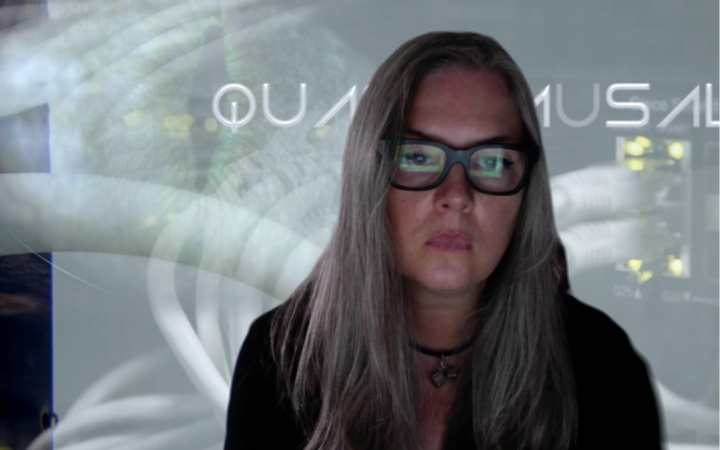 Ami Clarke, a woman with long hair and glasses with a thick, square frame, looks at a screen. This screen is reflected in her glasses.