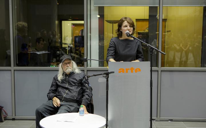 Links im Bild sitzt Ira Schneider, Medienkünstler, auf einem Stuhl und hört Daria Mille, Kuratorin am ZKM, bei ihrem Vortrag zu.
