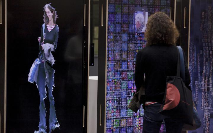 Eine braunhaarige Frau steht vor der Kunstinstallation YOU:RE CODE und sieht ihre digitale Spiegelung.