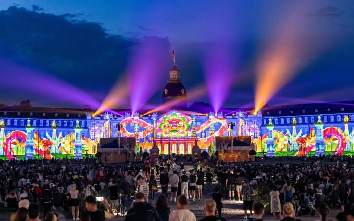 The façade of the baroque palace in Karlsruhe can be seen. The winning entry of the BBBank Award 2023 "BhinnekaExpress", 2023, The Fox, The Folks is projected. The castle glows in bright colours and laser beams shine into the sky. 